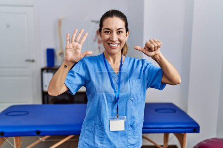 Foto de Young hispanic woman wearing physiotherapist uniform standing at clinic showing and pointing up with fingers number six while smiling confident and happy. - Imagen libre de derechos