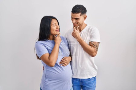Photo for Young hispanic couple expecting a baby standing over background looking confident at the camera smiling with crossed arms and hand raised on chin. thinking positive. - Royalty Free Image