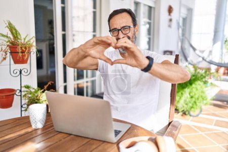 Foto de Hombre de mediana edad usando computadora portátil en casa sonriendo en el amor haciendo forma de símbolo del corazón con las manos. concepto romántico. - Imagen libre de derechos