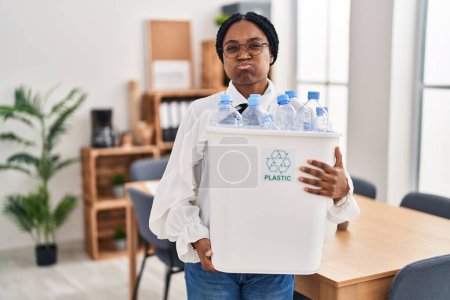 Photo for African american woman working at the office holding plastic bottle for recycling puffing cheeks with funny face. mouth inflated with air, catching air. - Royalty Free Image