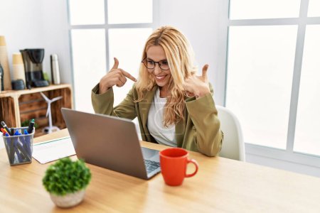 Foto de Beautiful blonde woman working at the office with laptop looking confident with smile on face, pointing oneself with fingers proud and happy. - Imagen libre de derechos