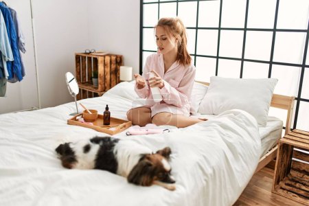 Téléchargez les photos : Jeune femme caucasienne appliquant un traitement de lotion de peau assis sur le lit avec chien à la chambre - en image libre de droit