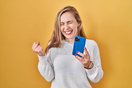 Photo for Young blonde woman using smartphone typing message very happy and excited doing winner gesture with arms raised, smiling and screaming for success. celebration concept. - Royalty Free Image