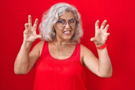 Foto de Mujer de mediana edad con el pelo gris de pie sobre fondo rojo sonriendo divertido haciendo gesto de garra como gato, expresión agresiva y sexy - Imagen libre de derechos