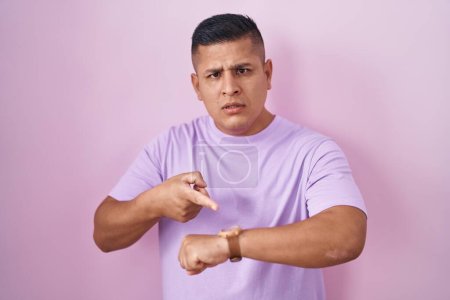 Foto de Joven hombre hispano de pie sobre fondo rosa con prisa apuntando a ver el tiempo, la impaciencia, molesto y enojado por retraso plazo - Imagen libre de derechos
