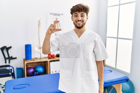 Téléchargez les photos : Jeune homme arabe travaillant à la clinique de récupération de la douleur geste souriant et confiant avec la main faisant signe de petite taille avec les doigts regardant et la caméra. concept de mesure. - en image libre de droit