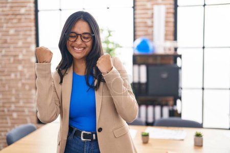 Photo for Brunette woman working at the office wearing glasses very happy and excited doing winner gesture with arms raised, smiling and screaming for success. celebration concept. - Royalty Free Image