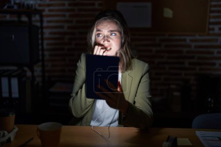 Téléchargez les photos : Femme caucasienne blonde travaillant au bureau la nuit à l'air stressée et nerveuse avec les mains sur les ongles mordants de la bouche. problème d'anxiété. - en image libre de droit