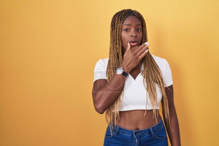 Photo for African american woman with braided hair standing over yellow background looking fascinated with disbelief, surprise and amazed expression with hands on chin - Royalty Free Image