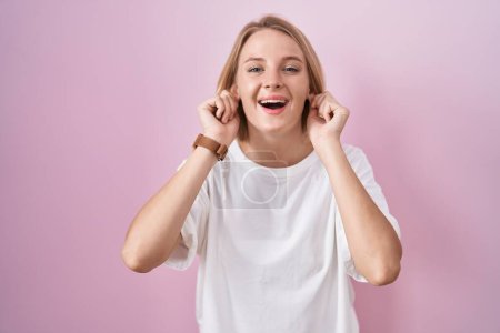 Photo for Young caucasian woman standing over pink background smiling pulling ears with fingers, funny gesture. audition problem - Royalty Free Image