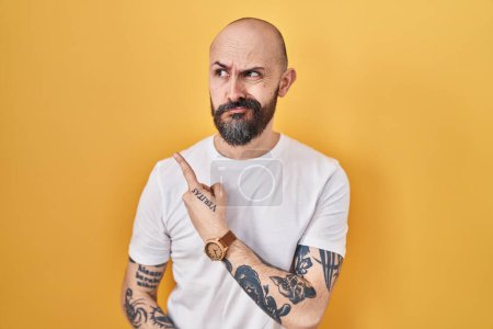 Foto de Joven hombre hispano con tatuajes de pie sobre fondo amarillo señalando a un lado preocupado y nervioso con el dedo índice, preocupado y sorprendido expresión - Imagen libre de derechos