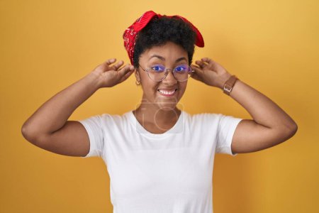 Foto de Mujer afroamericana joven de pie sobre fondo amarillo sonriendo tirando de las orejas con los dedos, gesto divertido. problema de audición - Imagen libre de derechos