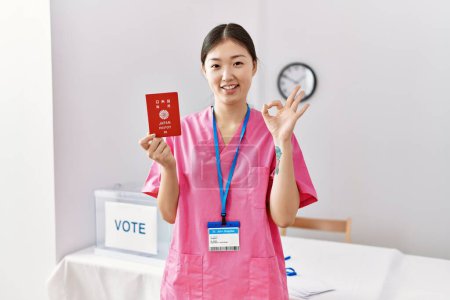 Téléchargez les photos : Jeune infirmière asiatique femme à l'élection de campagne politique tenant passeport japonais faire ok signe avec les doigts, sourire geste amical excellent symbole - en image libre de droit