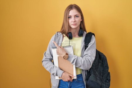 Téléchargez les photos : Jeune femme caucasienne portant un sac à dos étudiant et tenant des livres pointant de côté inquiet et nerveux avec index, expression préoccupée et surprise - en image libre de droit