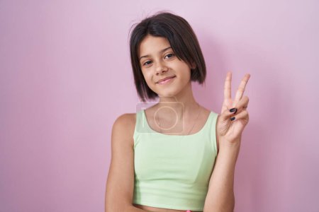 Téléchargez les photos : Jeune fille debout sur fond rose souriant avec le visage heureux clin d'oeil à la caméra faisant signe de victoire avec les doigts. numéro deux. - en image libre de droit