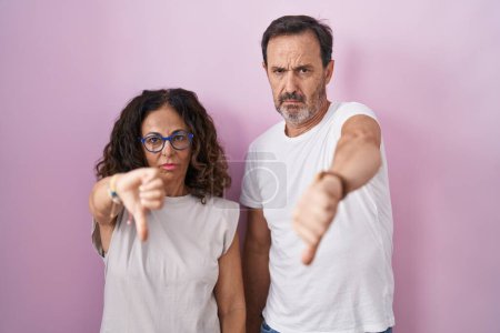 Photo for Middle age hispanic couple together over pink background looking unhappy and angry showing rejection and negative with thumbs down gesture. bad expression. - Royalty Free Image