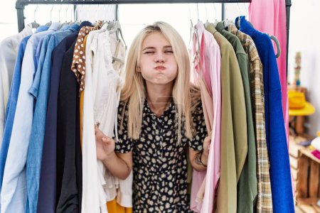 Photo for Young blonde woman searching clothes on clothing rack puffing cheeks with funny face. mouth inflated with air, catching air. - Royalty Free Image