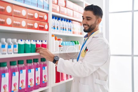 Téléchargez les photos : Jeune homme arabe pharmacien tenant la médecine sur les étagères à la pharmacie - en image libre de droit