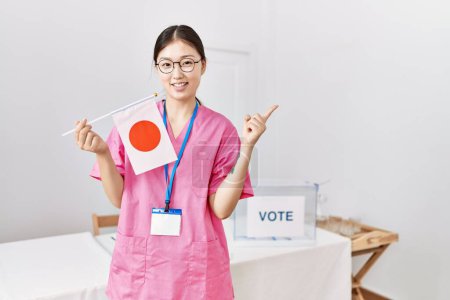 Téléchargez les photos : Jeune infirmière asiatique femme à l'élection de campagne politique tenant drapeau japonais souriant heureux pointant avec la main et le doigt vers le côté - en image libre de droit