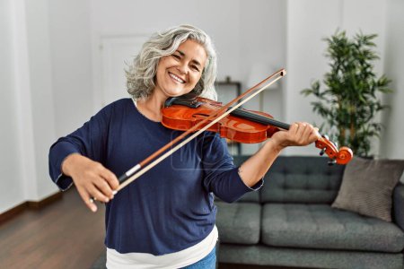 Photo for Middle age grey-haired artist woman playing violin standing at home. - Royalty Free Image