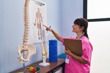 Photo for Young beautiful hispanic woman physiotherapist reading document looking anatomical model at rehab clinic - Royalty Free Image