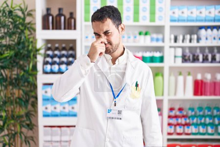 Foto de Handsome hispanic man working at pharmacy drugstore smelling something stinky and disgusting, intolerable smell, holding breath with fingers on nose. bad smell - Imagen libre de derechos