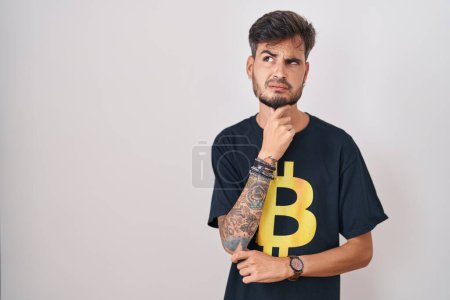 Téléchargez les photos : Jeune homme hispanique avec des tatouages portant Bitcoin t-shirt penser inquiet d'une question, préoccupé et nerveux avec la main sur le menton - en image libre de droit