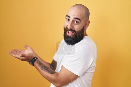 Foto de Joven hombre hispano con barba y tatuajes de pie sobre fondo amarillo señalando a un lado con las manos abiertas las palmas mostrando espacio de copia, presentando anuncio sonriendo emocionado feliz - Imagen libre de derechos