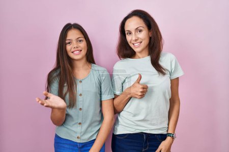 Téléchargez les photos : Jeune mère et fille debout sur fond rose montrant la main de la paume et faisant un geste correct avec les pouces levés, souriant heureux et joyeux - en image libre de droit