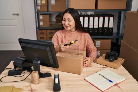 Téléchargez les photos : Jeune femme hispanique ecommerce travailleur d'affaires déballage boîte en carton en utilisant cutter au bureau - en image libre de droit