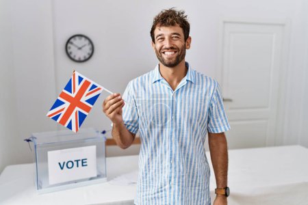 Foto de Joven hombre guapo en las elecciones de campaña política sosteniendo bandera del Reino Unido mirando positiva y feliz de pie y sonriendo con una sonrisa confiada mostrando los dientes - Imagen libre de derechos