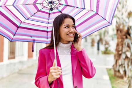 Foto de Mujer latina joven hablando en el teléfono inteligente con paraguas en la calle - Imagen libre de derechos