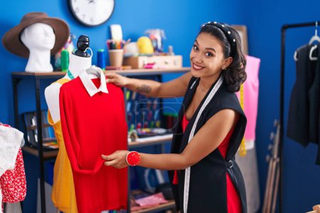 Téléchargez les photos : Jeune belle femme hispanique tailleur souriant confiant tenant t-shirt à l'usine de vêtements - en image libre de droit