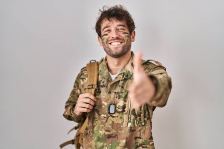 Foto de Joven hispano vistiendo uniforme camuflaje ejército sonriente amistoso ofreciendo apretón de manos como saludo y bienvenida. negocios exitosos. - Imagen libre de derechos