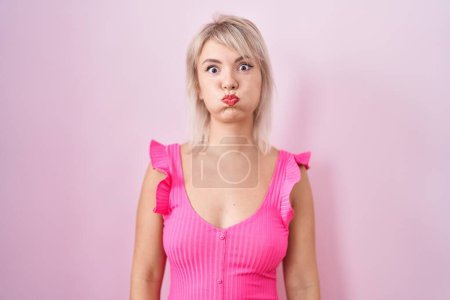 Foto de Joven mujer caucásica de pie sobre fondo rosa hinchando mejillas con cara divertida. boca hinchada de aire, expresión loca. - Imagen libre de derechos