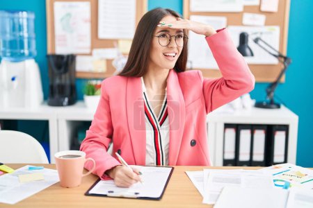 Photo for Young hispanic woman working at the office wearing glasses very happy and smiling looking far away with hand over head. searching concept. - Royalty Free Image