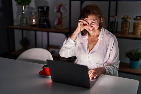 Foto de Mujer hispana de mediana edad usando portátil en casa por la noche haciendo un gesto bien con la mano sonriente, los ojos mirando a través de los dedos con la cara feliz. - Imagen libre de derechos