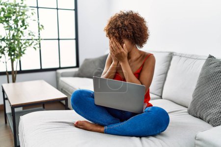 Photo for Young african american woman sitting on the sofa at home using laptop with sad expression covering face with hands while crying. depression concept. - Royalty Free Image