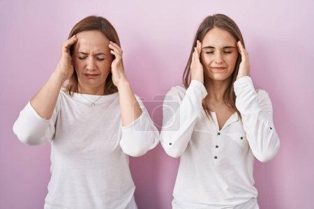 Foto de Madre de mediana edad y joven hija de pie sobre fondo rosa con la mano en la cabeza, dolor de cabeza porque el estrés. padecer migraña. - Imagen libre de derechos