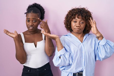 Téléchargez les photos : Two african women standing over pink background confused and annoyed with open palm showing copy space and pointing finger to forehead. think about it. - en image libre de droit