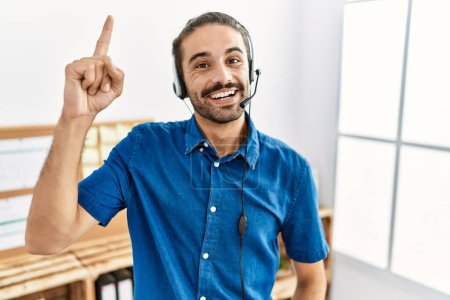 Foto de Young hispanic man with beard wearing call center agent headset at the office pointing finger up with successful idea. exited and happy. number one. - Imagen libre de derechos