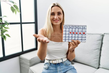 Young blonde woman holding heart calendar smiling cheerful offering palm hand giving assistance and acceptance. 