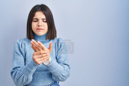Foto de Mujer hispana joven de pie sobre fondo azul sufriendo dolor en manos y dedos, inflamación de la artritis - Imagen libre de derechos