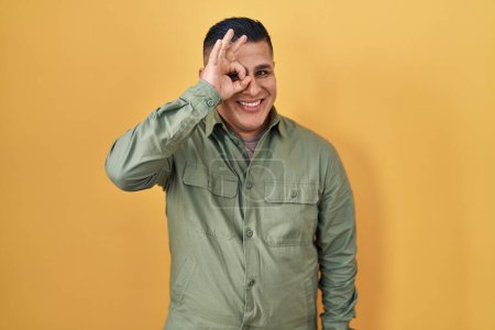 Téléchargez les photos : Jeune homme hispanique debout sur fond jaune faisant un geste correct avec la main souriante, les yeux regardant à travers les doigts avec un visage heureux. - en image libre de droit