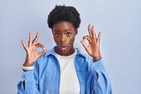 Foto de Mujer afroamericana de pie sobre fondo azul mirando sorprendido y sorprendido haciendo símbolo de aprobación bien con los dedos. expresión loca - Imagen libre de derechos