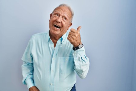 Foto de Hombre mayor con el pelo gris de pie sobre fondo azul sonriendo con la cara feliz mirando y señalando a un lado con el pulgar hacia arriba. - Imagen libre de derechos