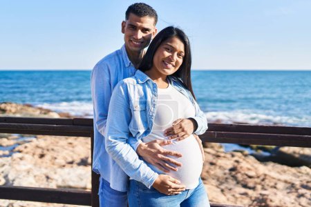 Photo for Young latin couple expecting baby hugging each other standing at seaside - Royalty Free Image