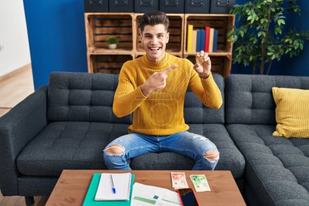 Téléchargez les photos : Jeune homme hispanique tenant Bitcoin argent virtuel souriant heureux pointant avec la main et le doigt - en image libre de droit