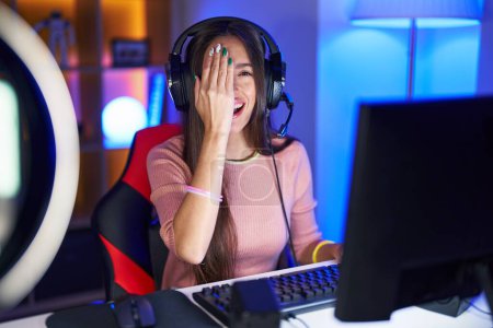 Téléchargez les photos : Jeune femme hispanique jouant à des jeux vidéo couvrant un oeil avec la main, sourire confiant sur le visage et l'émotion surprise. - en image libre de droit