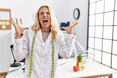 Photo for Beautiful blonde nutritionist woman at dietitian clinic crazy and mad shouting and yelling with aggressive expression and arms raised. frustration concept. - Royalty Free Image